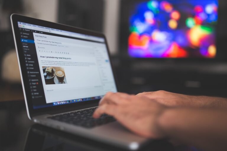 Hands typing on a laptop with a blog post visible, cozy indoor setting with colorful screen in background.